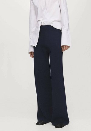 Persona che indossa pantaloni navy a vita alta e gamba larga abbinati a una camicia bianca a maniche lunghe e scarpe nere su uno sfondo neutro.