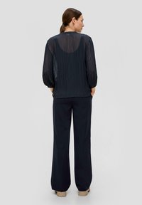 Marineblauwe geplooide blouse met een doorzichtige achterkant, lange pofmouwen, gecombineerd met ruime zwarte broeken en lichtbeige schoenen.