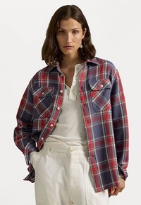 Red and navy plaid shirt with button-down front, two chest pockets, and a relaxed fit, paired with a light-colored top and cream pants.