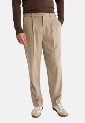 Pantaloni plissettati beige realizzati in tessuto morbido con una vestibilità comoda, dotati di chiusura con bottone e tasche laterali, abbinati a sneaker bianche.