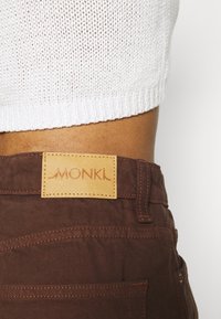 Los pantalones marrones presentan un parche de cuero beige con "MONKI" en relieve. Las costuras visibles y los detalles de los bolsillos realzan el diseño. Se muestra un top de punto blanco de corte corto.