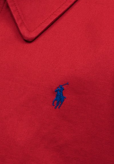 Κόκκινο ύφασμα με μπλε κεντημένο λογότυπο παίκτη πόλο πάνω σε άλογο, με λεπτομερείς ραφές ορατές κοντά στην περιοχή του γιακά.