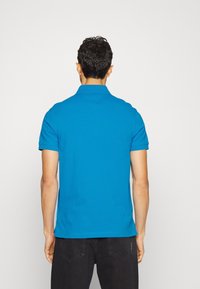 Muž v jasně modré polokošili s krátkým rukávem a černých džínech, viděn zezadu na prostím světlém pozadí.