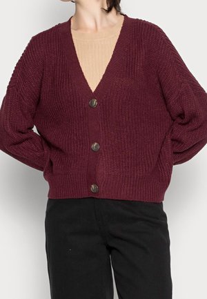 Personne portant un cardigan en maille bordeaux boutonné sur un haut côtelé beige et un pantalon noir, debout les mains derrière le dos.