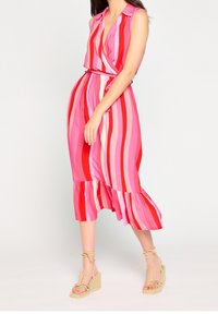 Bezramionowa sukienka w różowo-czerwone paski o kroju kopertowym, z marszczonym wcięciem w talii i falbaniastym dołem. W połączeniu z beżowymi sandałami na koturnie.