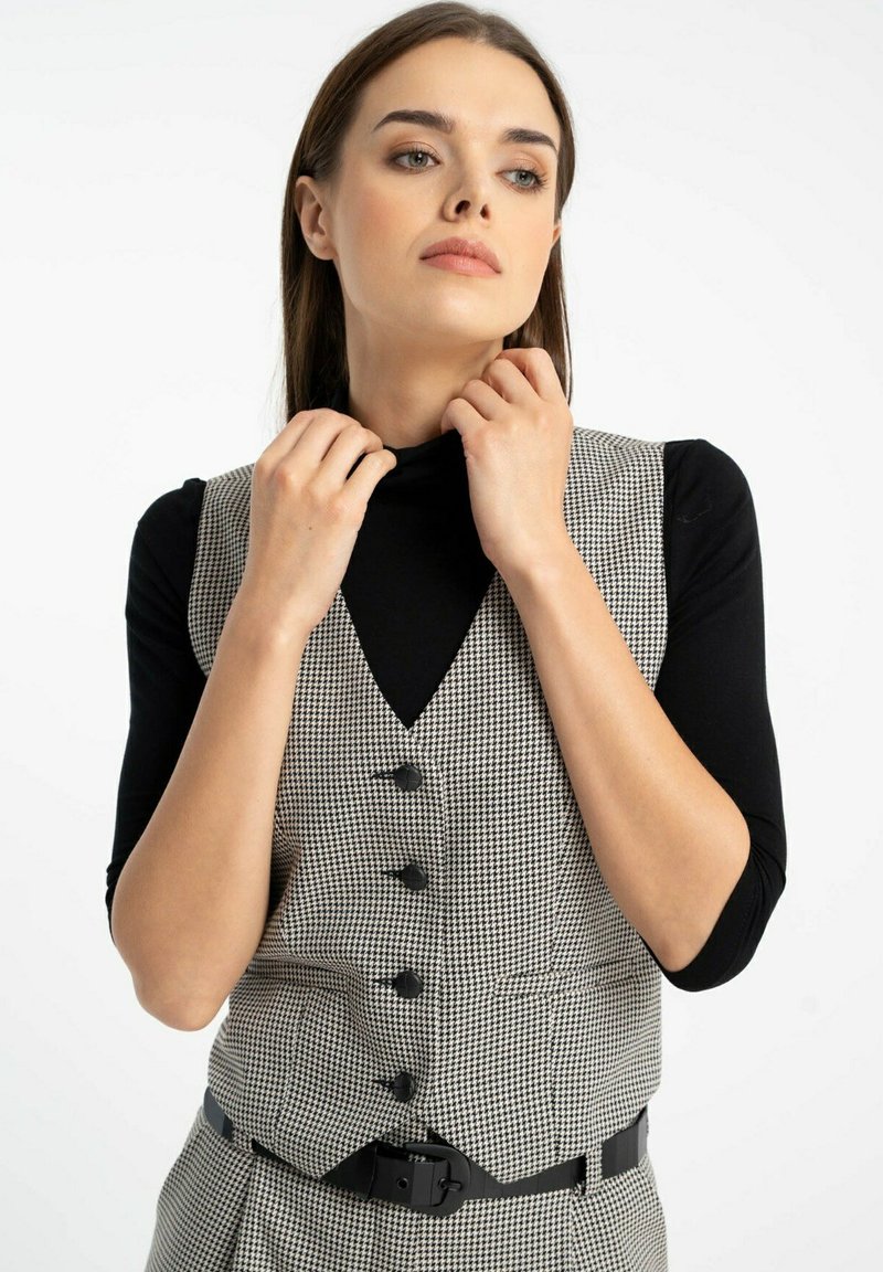 Kobieta poprawiająca kołnierzyk czarnej bluzki noszonej pod kamizelką w czarno-białą kratkę z przodu zapinaną na guziki, w połączeniu z dopasowanymi spodniami.