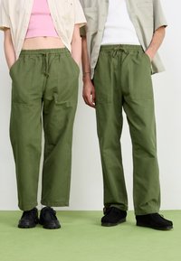 Pantalons en coton verts avec taille élastique et poches, associés à des chaussures noires. Le tissu possède une texture lisse et mate.