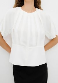 Femme portant un chemisier blanc à manches courtes plissé avec un col rond et une jupe noire, debout les mains derrière le dos.