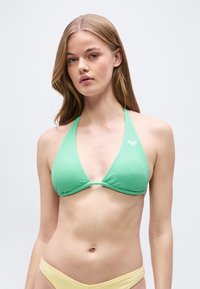 Νεαρή γυναίκα που φοράει πράσινο πλεκτό μπικίνι με στυλ halter και ανοιχτό κίτρινο κατώτερο μέρος μπικίνι, στέκεται μπροστά σε λευκό φόντο.