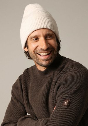 Pull marron à côtes avec un col haut, porté avec un bonnet blanc. Le pull présente une étiquette marron sur la manche gauche.