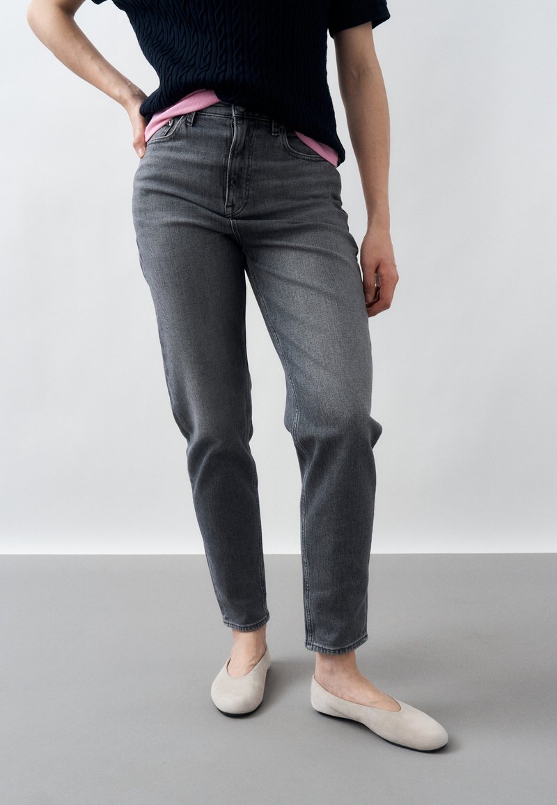 Person wearing high-waisted gray jeans, black short-sleeve sweater layered over pink shirt, and light beige flat shoes, standing with one hand on hip.