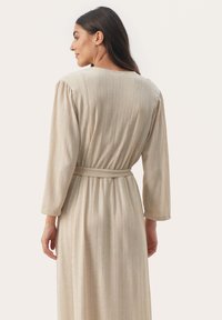 Langes, hellbeiges Kleid mit vertikaler gerippter Textur, langen Ärmeln und einem betonten Taillengürtel. Offener Rücken mit einem runden Ausschnitt.
