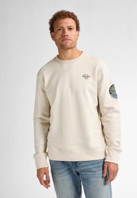 Felpa beige con scollo rotondo e polsini a costine, caratterizzata da una stampa sul lato sinistro del petto e un logo ricamato sulla manica destra.