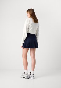 Hvit langermet topp kombinert med et marineblått plisse skjørt. Modellen har på seg hvite sokker og chunky sneakers med svarte detaljer.