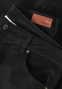 Čierne rifle z denimu s hladkou textúrou, vybavené hnedou koženou značkou s nápisom „TATUUM“ a kovovými doplnkami v páse a na vrecku.