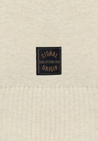 Material tricotat de culoare crem, cu textură cu nervuri, având o etichetă rectangulară neagră pe care este scris "SIGNAL ORIGIN QUALITY SINCE 1967" cu litere aurii.