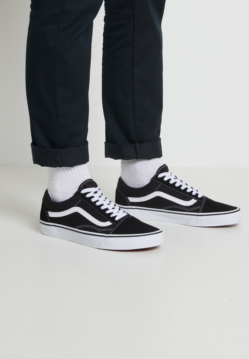 Personne portant un pantalon noir et des chaussettes blanches avec des baskets basses noires et blanches debout sur un sol clair.