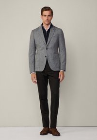 Blazer gris texturizado con cierre de dos botones, combinado con una camisa de cremallera azul marino y pantalones oscuros, completando el atuendo con botas marrones.