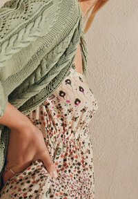 Frau in einem grünen gestrickten Schal über einem Blumenkleid, die Hand auf der Taille des Kleides ruhend, nahe einer strukturierten beigen Wand.