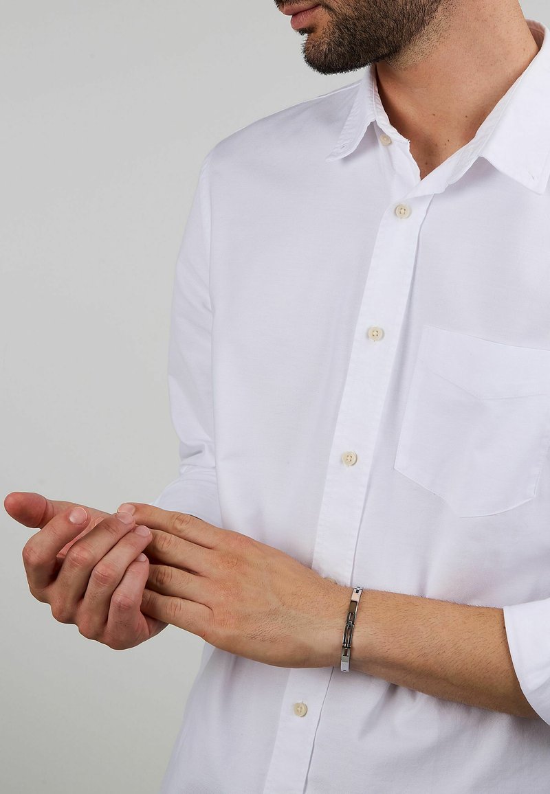 Bracciale in metallo argento su un polso, caratterizzato da un design a catena. L'individuo indossa una camicia bianca con bottoni dotata di una tasca sul petto.