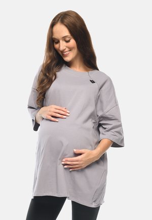 Schwangere Frau in lockerem grauem Oberteil hält sanft ihren Bauch, lächelt und blickt nach unten, steht vor einem schlichten hellen Hintergrund.