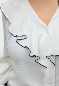 Blouse blanche avec un décolleté en V, ornée d'une finition à volants soulignée en noir. Tissu texturé et fermeture à un bouton à l'avant.