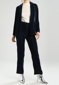 Námořnický sametový blazer se dvěma předními kapsami, v kombinaci s odpovídajícími sametovými kalhotami a krémovým žebrovaným rolákem, doplněný stříbrnými botami na podpatku.