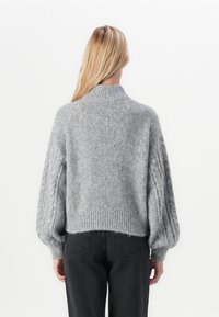 Frau mit langen blonden Haaren, die einen grauen strukturierten Strickpullover mit Puffärmel und schwarze hochtaillierte Jeans trägt und sich von hinten zeigt.