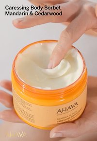 Orange plastburk med kräm med en vit, krämig konsistens. En hand dyker ner i burken. Etiketten innehåller "AHAVA" och produktinformation.