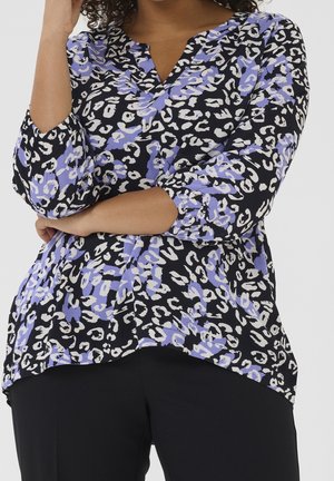 Woman wearing a black blouse with white and lavender leopard print and three-quarter sleeves, arms crossed, paired with black pants.