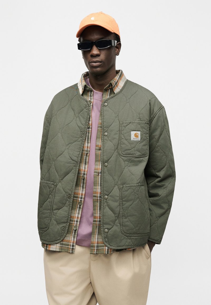 Homme portant une casquette orange, des lunettes de soleil noires, une veste matelassée verte sur une chemise à carreaux et un t-shirt rose, avec un pantalon beige, debout devant un fond blanc.