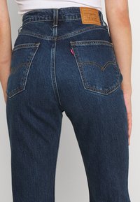 Tmavě modré džíny z denimu s vysokým pasem, dvěma zadními kapsami s prošitými detaily a hnědou koženou etiketou na pase.