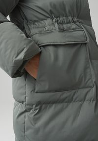 Giacca imbottita di un verde tenue con tasca frontale, vita elastica e texture liscia per calore e comfort.