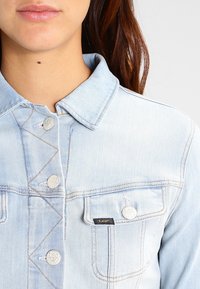 Hellblaues Jeansjacke mit klassischem Kragen, silbernen Druckknöpfen, einer Brusttasche mit schwarzem Etikett und sichtbaren Nahtdetails.