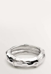 Pulsera de metal plateado con textura martillada, que presenta una forma redondeada y ligeramente irregular y una superficie lisa y reflectante.