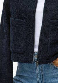 Marinblå kort fleecejacka med två framfickor, en silverfärgad dragkedja och en mjuk textur. Bärs över en vit topp och blå högmidjade jeans.