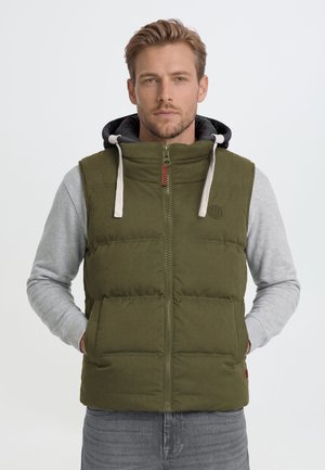 Homme portant une veste matelassée vert olive avec fermeture éclair à l'avant et un sweat à capuche gris en dessous, debout devant un fond clair uni.