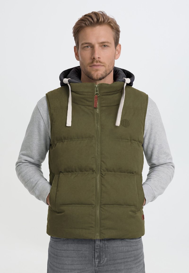 Hombre con un chaleco acolchado verde oliva con cremallera frontal y sudadera gris con capucha debajo, de pie frente a un fondo claro y sencillo.