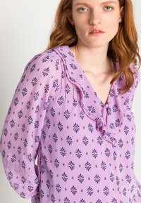 Blusa lavanda feita de um tecido leve, apresentando um decote com babados, mangas longas e padrões florais escuros por toda a peça.