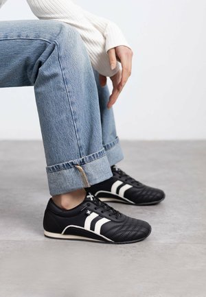 Zapatos negros de cordones con una parte superior de cuero texturizado, rayas diagonales blancas y una silueta de bajo perfil. Llevados con unos vaqueros azules ajustados.
