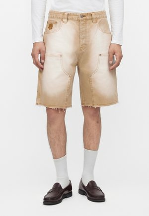 Mand iført beige cargoshorts med rå kanter, hvide sokker, mørkebrune loafers og en langærmet hvid skjorte, der står mod en hvid baggrund.