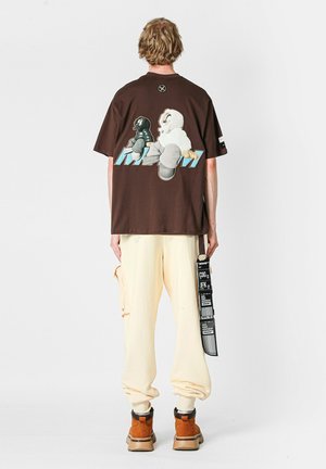 Camiseta grande de color marrón con arte gráfico de dos personajes en la parte trasera. Combinada con pantalones amarillos y botas marrones, mostrando diferentes texturas.