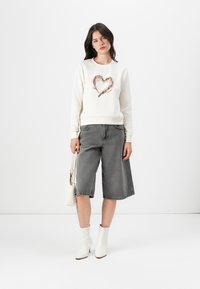 Lys kremfarget sweatshirt med et paljettdekorert hjerte, kombinert med grå denim culotte-bukser, hvite ankelstøvler og en liten hvit håndveske.