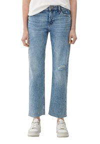 Mujer vistiendo jeans rectos desgastados de color azul claro y zapatillas blancas, mostrada de la cintura a los pies sobre un fondo blanco.