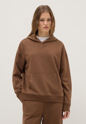 Jeune femme portant un sweat à capuche marron et un pantalon assorti, debout sur un fond clair uni avec une expression neutre.