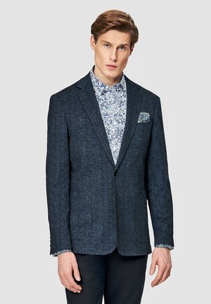 Mężczyzna w ciemnoniebieskiej marynarce na białej koszuli z niebieskim kwiatowym wzorem, stojący na tle jednolitego jasnoszarego tła.