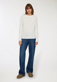 Kremowy sweter o prążkowanej fakturze z okrągłym dekoltem i długimi rękawami, zestawiony z szerokimi niebieskimi dżinsami i brązowymi butami wsuwanymi.
