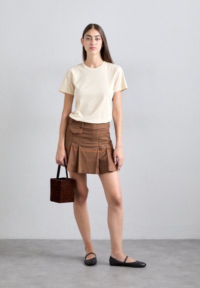 Beige cotton t-shirt, brown plaid pleated skirt with orange lines, black flat shoes, small brown bag, standing on gray background.