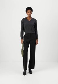 Maglione grigio a collo a V con maniche lunghe abbinato a pantaloni neri sartoriali. Il modello tiene una borsa verde indossando stivali neri alla caviglia.