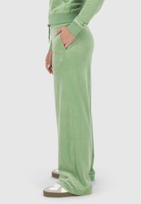 Joggers en velours vert clair avec poches latérales, taille élastique et cordon de serrage. Texture lisse avec un branding subtil sur la cuisse.